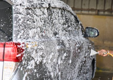Biri araba yıkamada gri bir aracı köpükle kaplamak için köpük püskürtücü kullanıyor. Parlak güneş ışığı baloncukları vurgular, ışıltılı bir etki yaratır..