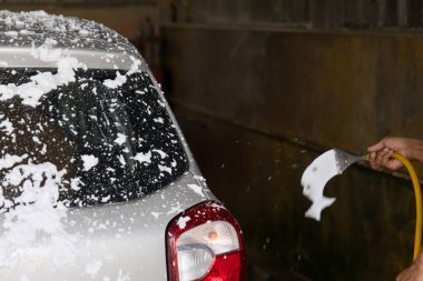 Akşamları garajda, bir insan gümüş bir arabanın sabununu temizlemek için kalın bir köpük tabakası uyguladıktan sonra durulamak için hortum kullanır..