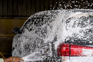 Gündüz vakti profesyonel bir araba yıkama alanında duran bir kişi, köpük aplikatör kullanarak bir aracı temizliyor, sabun ve su uyguluyor..
