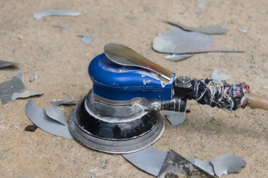 Mavi bir zımparalama aracı, dağınık metal parçalarıyla çevrili tozlu bir yüzeyde duruyor. Bu da el yapımı bir atölye projesinin sonucunu gösteriyor..