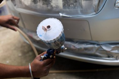 A person operates a spray gun while painting the front bumper of a car in an auto repair shop. The repair involves detailing and refinishing work to enhance the vehicle's appearance.