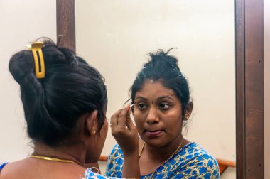 Güney Asyalı bir kadın makyaj yapıyor, aynadaki yansımasına dikkatlice bakıyor, güne hazırlanıyor..