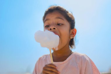 A young girl enjoys a sweet treat on a sunny day, biting into a large fluffy cloud of cotton candy against a bright blue sky. clipart