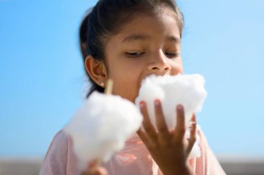 Girl joyfully eating cotton candy outdoors on a sunny day. Sweet treat, summer fun! clipart