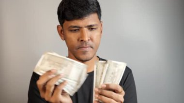 Siyah gömlekli bir adamın portresi banknotları inceliyor. Odaklanmış ifadesi finansal planlama veya bütçe öneriyor. Tarafsız arka plan konsantrasyonunu vurguluyor..