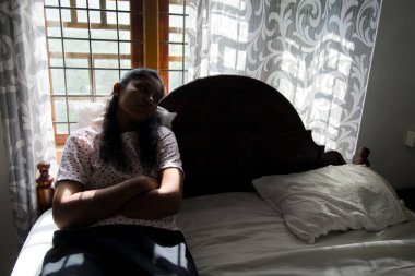 Asyalı bir kadının huzurlu bir görüntüsü. Düşünceli bir şekilde yatağında oturmuş pencereden dışarıyı seyrediyor..