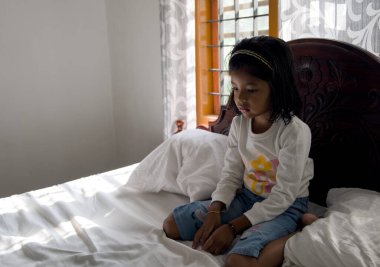 Küçük bir kızın sabah güneşinde yatağında oturduğu seçici odak görüntüsü. Üzgün görünüyor.