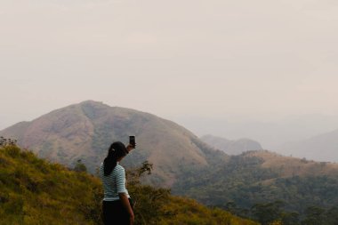 Bir kadın bir dağın zirvesinde duruyor, cep telefonuyla nefes kesen güneşin doğuşunu yakalıyor. Panoramik manzarayı seviyor.