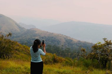 Genç bir kadın dağın zirvesinde duruyor, telefon kamerasıyla nefes kesici bir doğa sahnesi çekiyor. O, günlük açık hava kıyafetleri giyer.