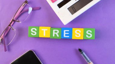 Renkli yapı taşları, kalem ve gözlüklerle birlikte mor bir arkaplanda stres kelimesini gösterir.