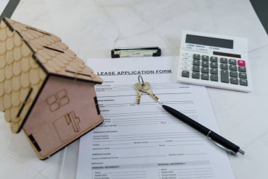 Kiralama başvuru formu, temiz bir yüzey üzerinde ahşap bir ev modeli, anahtarlar ve hesap makinesi ile birlikte doldurulur..