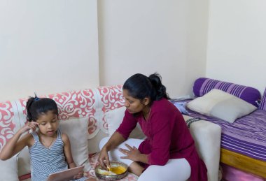Bir anne sıcak bir odada yemek pişirirken kızı tabletle meşgul oluyor, aile etkileşimlerini ve günlük yaşamını gösteriyor..