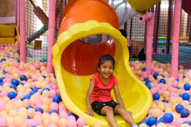 Neşeli bir çocuk, köpük toplarıyla dolu canlı bir oyun bahçesinde kayıyor, hoş bir şekilde gülümsüyor..