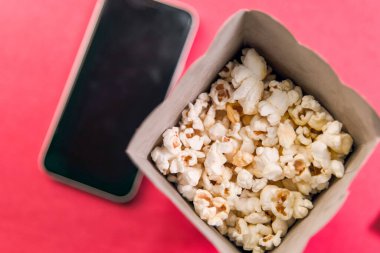 Bir kutu patlamış mısır canlı bir arka planda, akıllı telefonun yanında duruyor. Film gecesi ve eğlence temaları için ideal..