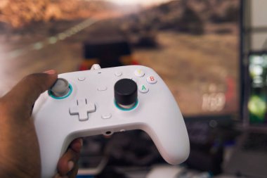 Bir el, kapalı bir oyun oturumu sırasında ekranda bir yarış oyunu gösterilirken bir beyaz oyun denetleyicisini kavrıyor.