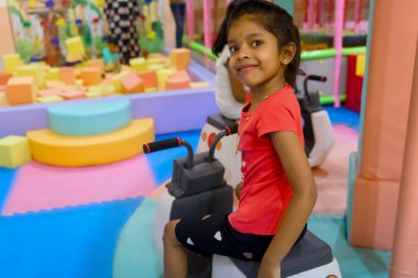 Genç kız, yumuşak bloklarla dolu, canlı bir oyun alanında mini bir oyuncağa binerken gülümsüyor..