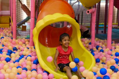 Çocuk, yumuşak toplarla dolu renkli bir oyun alanında sarı bir kaydıraktan kaymayı seviyor..