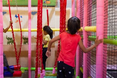 Çocuklar canlı kapalı bir oyun bahçesinde, tırmanma çerçeveleri ve renkli ağlarla eğlenceli aktivitelere girişiyor..