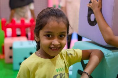 Genç kız, çocuklarla ve eğlence ekipmanlarıyla dolu canlı bir oyun alanında yumuşak bloklarla oynarken gülümsüyor..