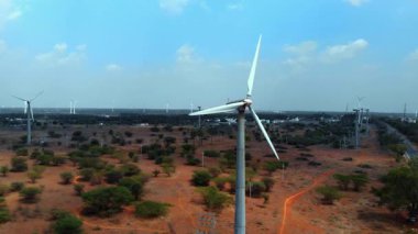 Rüzgâr türbinleri, kurak bir kırsal alanda, açık mavi gökyüzünün altında, seyrek bitki örtüsü ve uzak tepelerde dimdik durur..