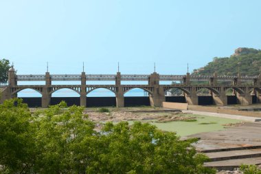 Açık mavi gökyüzünün altında yeşil ve tepelerle çevrili sakin bir su kütlesi üzerinde beton bir köprü uzanır..