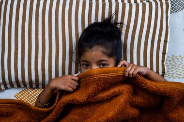 Bir çocuk, sıcak bir battaniyeden çizgili yastıklara sarılmış bir yatağa bakar ve rahat bir hava yaratır..