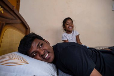 Bir baba, kızı arkasında mutlu bir şekilde oynarken yatakta dinlenir ve sıcak bir aile ortamı yaratır..