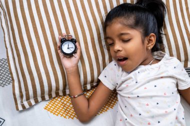 Genç kız çizgili bir yastıkta dinleniyor, rahat bir yatak odasında mışıl mışıl uyurken saati nazikçe tutuyor..