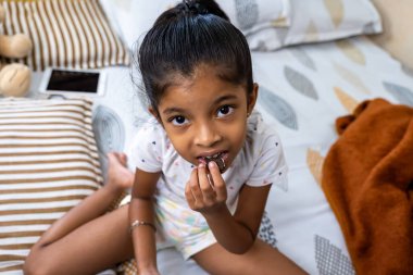 Genç bir kız, sıcak bir yatak odasında, yumuşak dokuları olan bir yatakta mutlu bir şekilde çikolata yiyor..