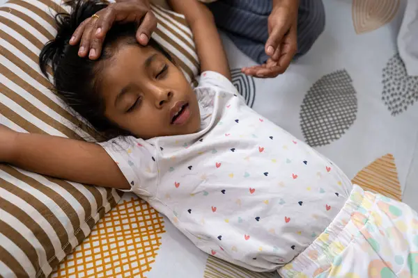 Genç bir kız sırtüstü yatıyor, gözleri kapalı, bir bakıcı nazikçe alnına dokunuyor..