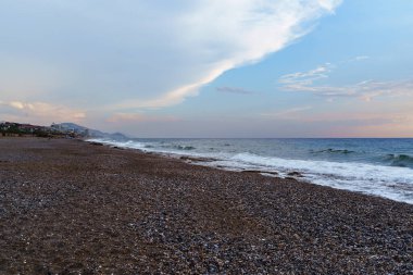 Gün batımında çakıl taşı plajının sakin bir sahnesi. Gökyüzü dramatik bulutlarla ve okyanus boyunca uzanan güneş ışınlarıyla dolu. Huzurlu ve rahatlatıcı bir atmosfer yaratıyorlar..