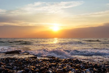 Okyanusun üzerinde güzel bir günbatımı ön planda parıldayan çakıl taşlarıyla doğal güzelliğin sakin ve sakin bir atmosferini yaratıyor..