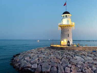 Alacakaranlıkta kayalık bir iskelede duran ışıklarla dekore edilmiş çarpıcı bir deniz feneri. Sakin deniz ve sakin gökyüzü şehrin etrafında huzurlu bir atmosfer yaratır..