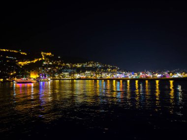 Deniz kıyısındaki bir şehrin göz kamaştırıcı gece manzarası, suya yansıyan parlak ışıklarla aydınlatılmış, resim gibi ve canlı bir atmosfer yaratıyor..