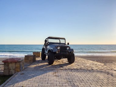 Gün batımında güzel bir plajın yanına park edilmiş engebeli bir off-road aracı. Okyanus dalgaları batan güneşin altında parlıyor, sakin ve maceracı bir sahne yaratıyor..