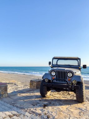Gün batımında güzel bir plajın yanına park edilmiş engebeli bir off-road aracı. Okyanus dalgaları batan güneşin altında parlıyor, sakin ve maceracı bir sahne yaratıyor..