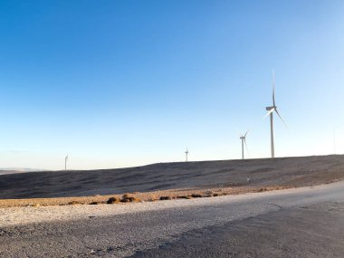 Bulutsuz mavi gökyüzünün altında ıssız ve çorak bir bataklıkta rüzgar türbinleriyle yenilenebilir enerji manzarası. Sürdürülebilirliği ve doğayla uyumu temsil eder.