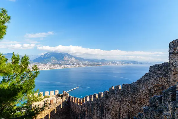 Sakin sularla dolu bir sahil şeridini gösteren nefes kesici bir manzara, tarihi kale duvarları ve görkemli dağlarla çerçevelenmiş, hepsi parlak ve berrak bir gökyüzü altında resim gibi ve sakin bir atmosfer yaratıyor..