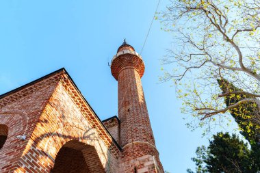 Kırmızı tuğladan yapılmış tarihi bir cami, kemerli bir galeri, ve uzun bir minare, doğal güneş ışığıyla yıkanmış ve ağaçlarla çevrili, dingin bir mimari ve kültürel ortam yaratıyor..