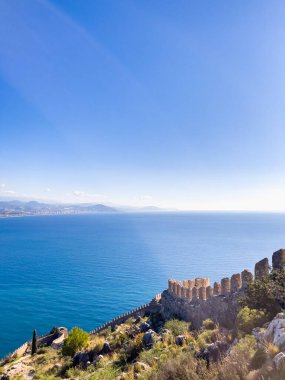 Canlı mavi sulara sahip sakin bir kıyı manzarası, arka planda dağlar, ve tarihi tahkimatlar, seyahat ya da doğa temaları için mükemmel.