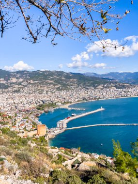 Antalya, Alanya şehrinin canlı bir limanı, etrafındaki dağları ve açık mavi gökyüzünü gösteren manzaralı bir görüntüsü. Seyahat ilhamları ve Akdeniz atmosferlerinin sergilenmesi için ideal.