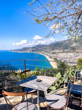Güzel bir sahil şehri ve canlı bir mavi okyanusa bakan manzaralı resimsel bir manzara. Sahne, parlak güneşli bir gökyüzünün altında, doğal ağaçlar ve yapraklar tarafından canlandırılmıştır..