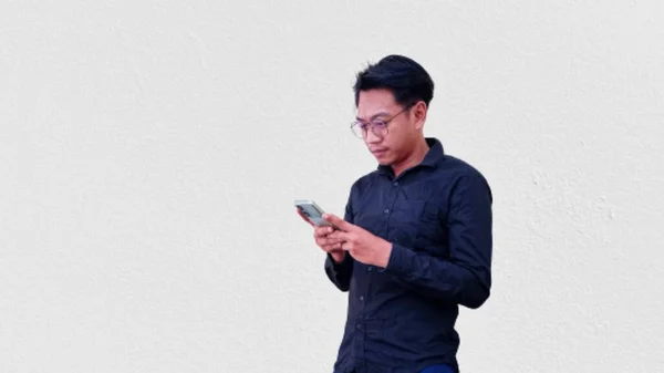 携帯電話を持っている黒いシャツを着た混乱したアジア人男性の肖像 白い背景に隔離された — ストック写真
