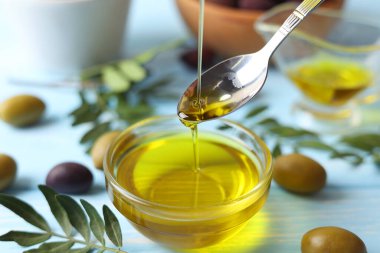 Gümüş bir kaşık altın zeytin yağını taze zeytin ve canlı yeşil yapraklarla çevrili bir kaseye serpiştirir.