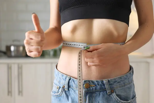 Bir kadın şık bir kot pantolon ve ekin bluzu giyerken belini mezurayla dikkatlice ölçer.