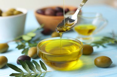 Gümüş bir kaşık altın zeytin yağını taze zeytin ve canlı yeşil yapraklarla çevrili bir kaseye serpiştirir.