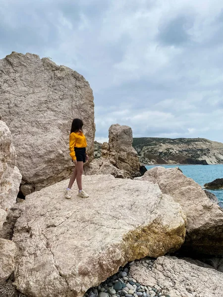Turuncu bluz ve şort giyen güzel kız bir kayanın üzerinde duruyor ve denize bakıyor, mavi suya. Yüksek kalite fotoğraf