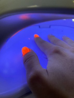 Mavi bir ultraviyole lambanın içinde neon turuncu manikürlü, jel cilası kurutan bir kadın el. Manikür süreci, kurutma aşaması. Yüksek kalite fotoğraf