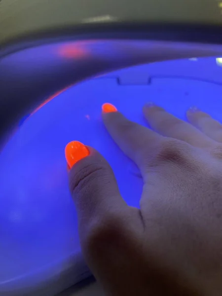 Mavi bir ultraviyole lambanın içinde neon turuncu manikürlü, jel cilası kurutan bir kadın el. Manikür süreci, kurutma aşaması. Yüksek kalite fotoğraf