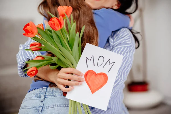Grateful mom hugging daughter girl, holding flowers bouquet, receiving hand drawn greeting card with loving heart from girl, smiling at camera. Mothers day, 8 march, concept. Head shot portrait. Close-up of hands holding a bouquet of tulips and a han